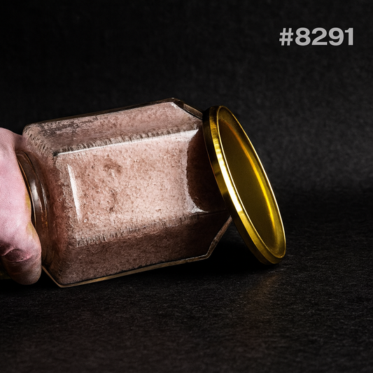 Close-up of a dusty square glass jar tipped on its side, with a golden lid leaning against it and a hand holding it; dark, moody setting.