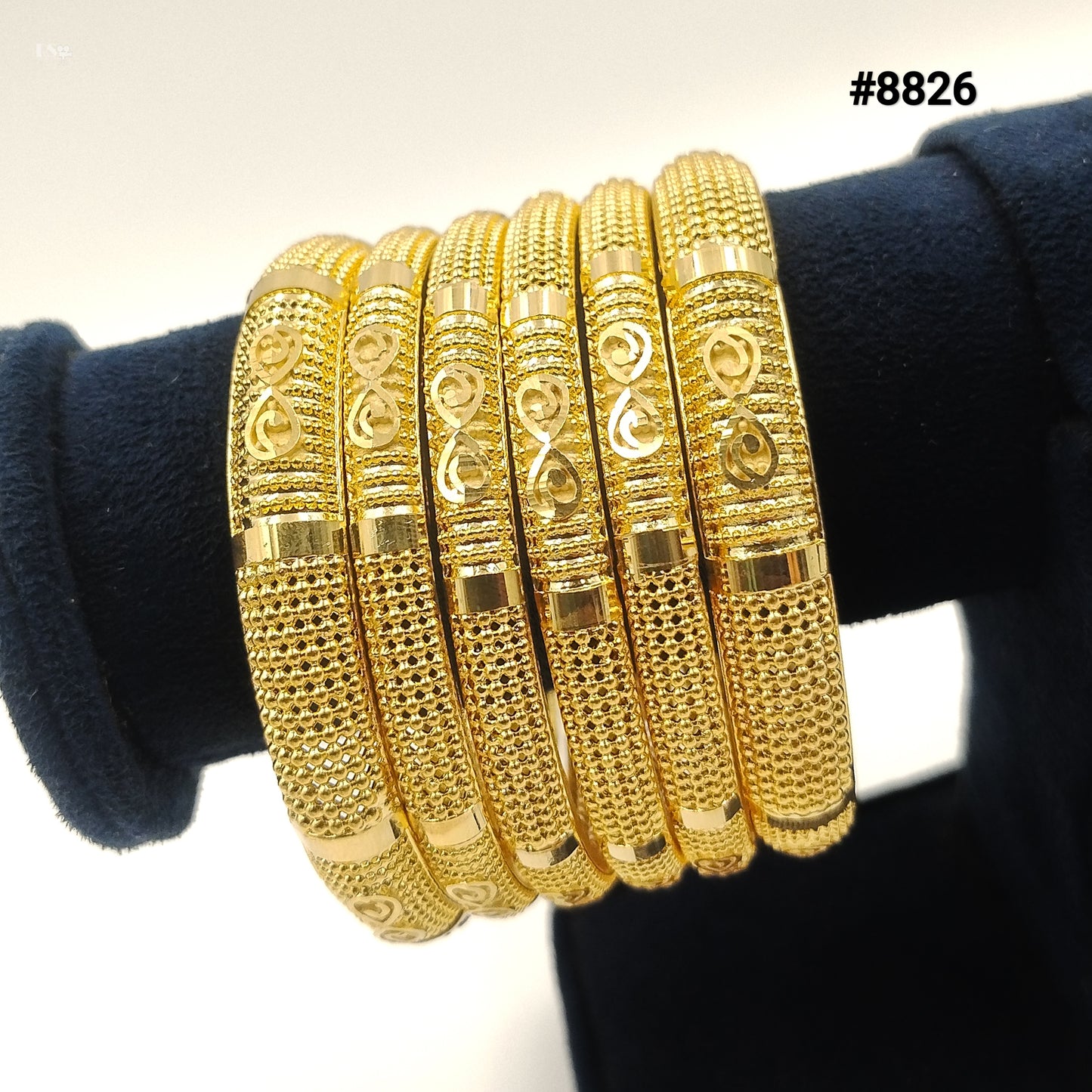 Six intricately designed gold bangles with textured and patterned details are displayed on a dark blue velvet stand. The item number #8826 is visible in the top right corner.