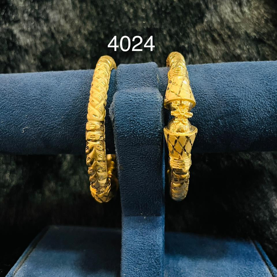 Two ornate gold bangles with engraved, textured designs rest on a dark blue velvet jewelry stand, labeled “4024” against a shadowy backdrop.