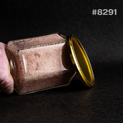 Close-up of a dusty square glass jar tipped on its side, with a golden lid leaning against it and a hand holding it; dark, moody setting.