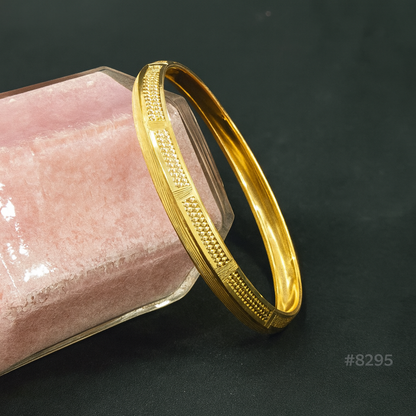 A gold bangle with textured and dotted patterns rests on a pink, hexagonal velvet jewelry box against a black background. The number #8295 appears in the lower right corner.