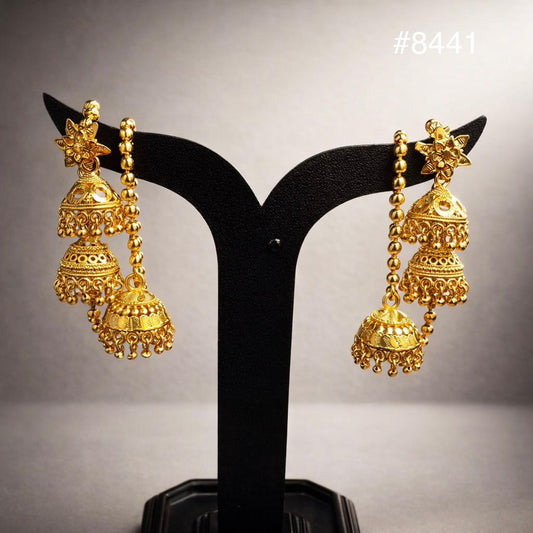 Ornate gold jhumka earrings with peacock details, chains, and tiered filigree bells displayed on a black stand; luxurious, traditional tone.
