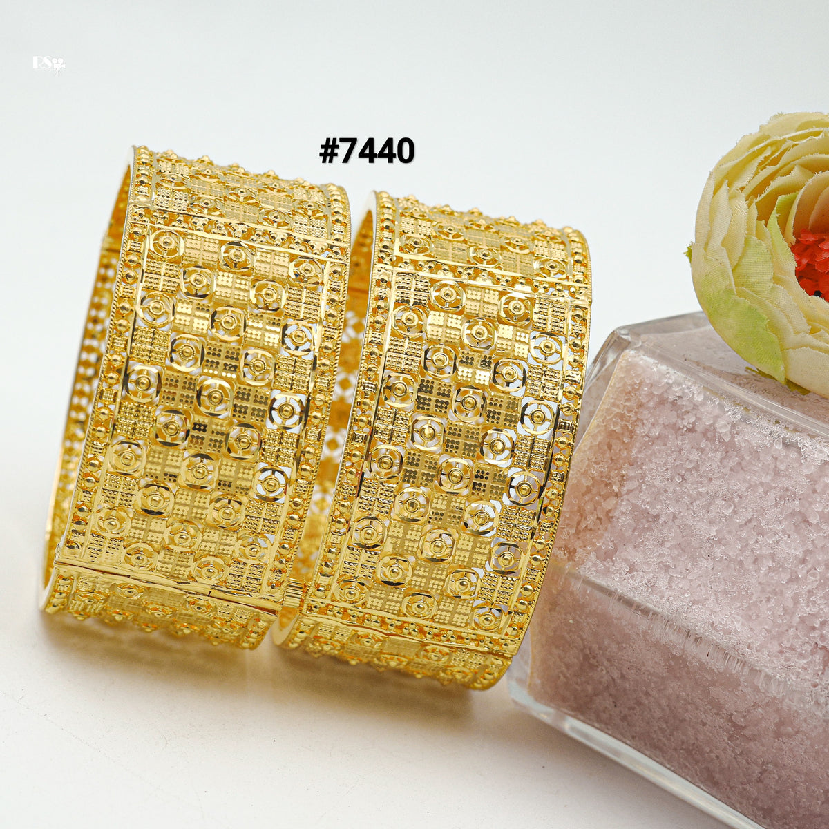 Pair of ornate gold bangles labeled #7440, featuring intricate geometric patterns, beside a pink textured block and pale yellow flower.