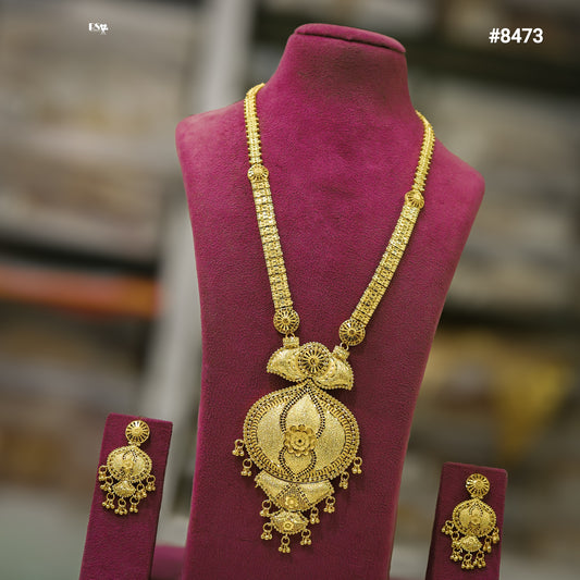 Ornate gold necklace with a large traditional pendant, flanked by matching earrings, displayed on a burgundy velvet bust; elegant jewelry showcase.