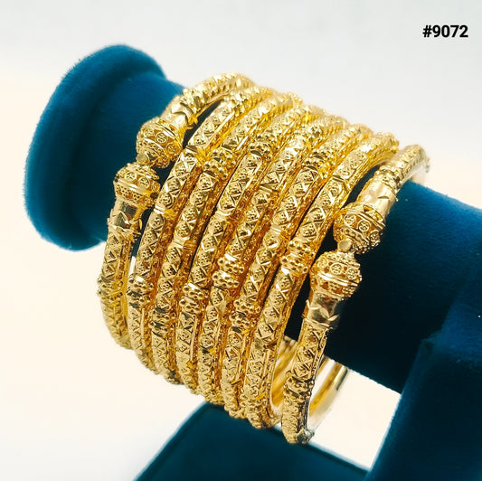 Close-up of stacked ornate gold bangles with engraved, beaded designs displayed on a dark teal velvet stand; luxurious jewelry, product #9072.